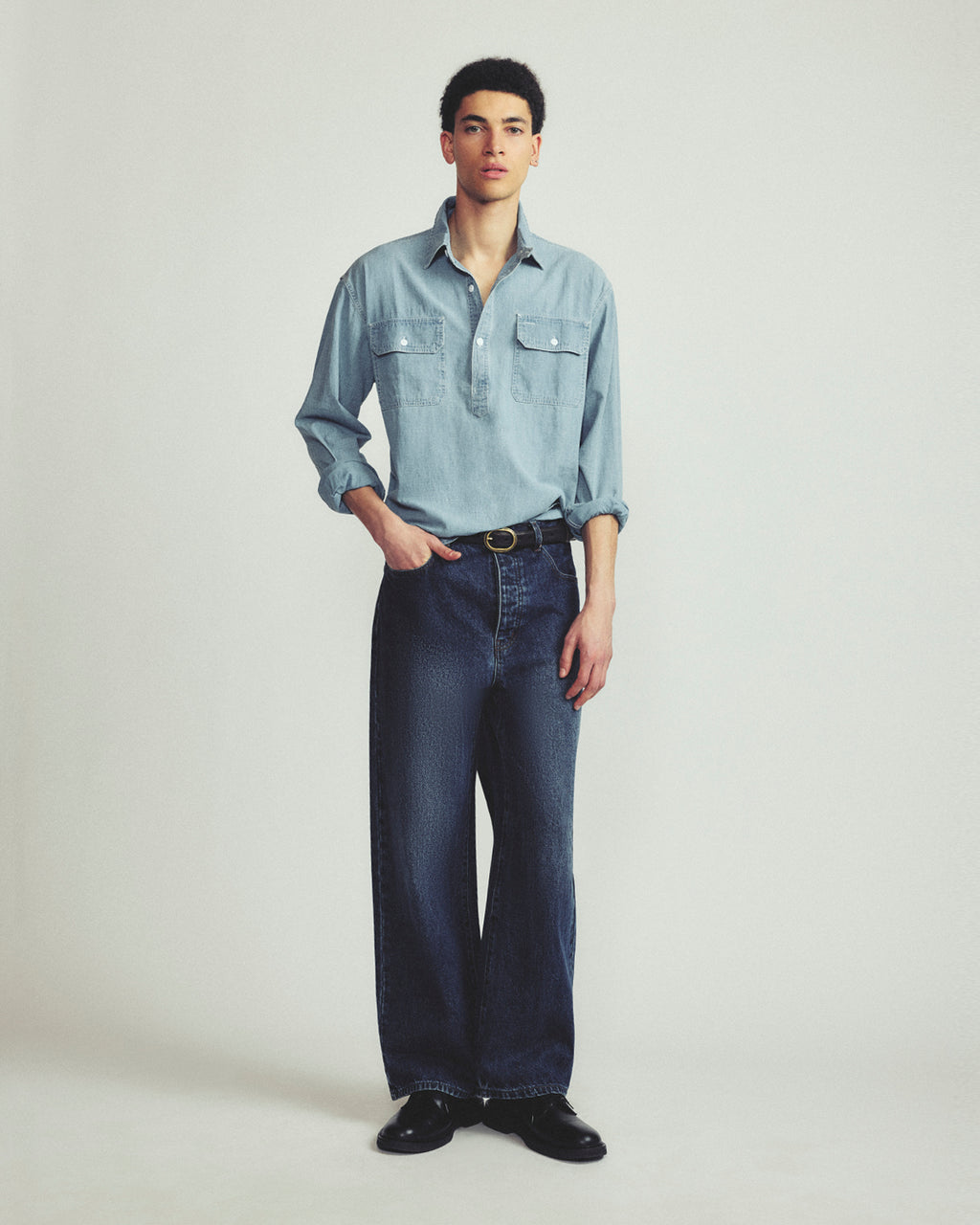 A male model stands against a light gray studio background wearing a light blue long-sleeve button-up shirt, dark blue wide-leg jeans, a black belt, and black shoes.