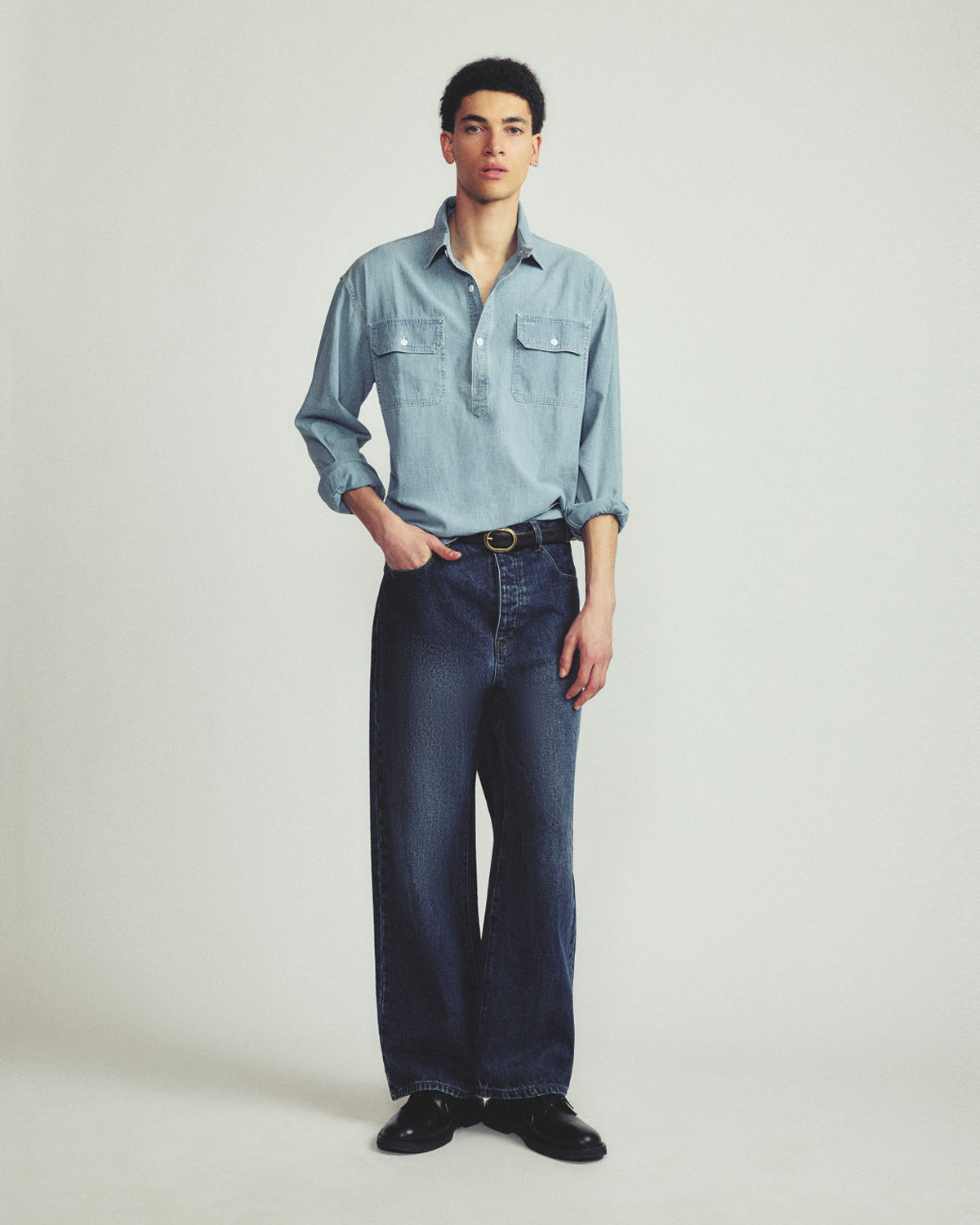 A male model stands against a light gray studio background wearing a light blue long-sleeve button-up shirt, dark blue wide-leg jeans, a black belt, and black shoes.