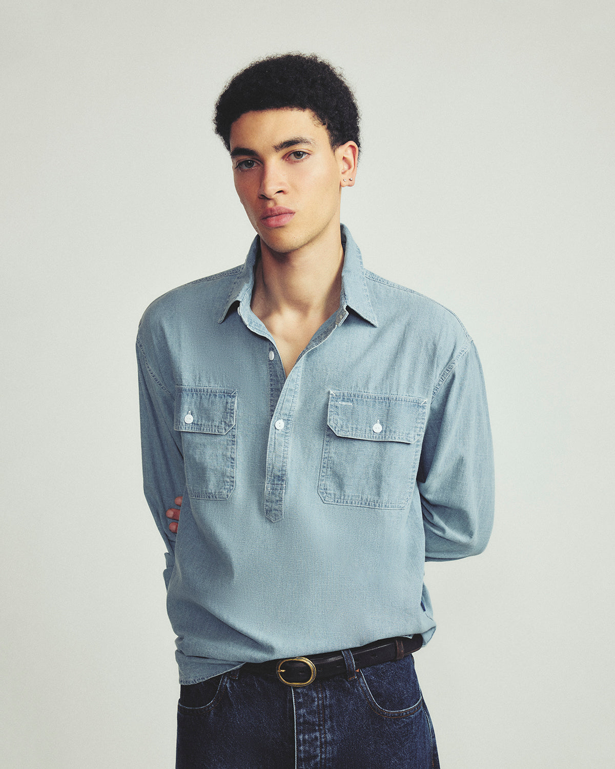 A young man wearing a light blue denim button-up shirt with two chest pockets and dark blue jeans, standing against a plain light gray studio background.